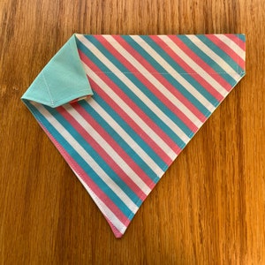 May include: A diagonally folded pet bandana with a mint green backing. The front has a pattern of alternating pink, white, and light blue stripes. The edges are neatly stitched, and the bandana is displayed on a wooden surface.