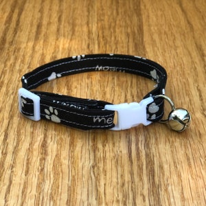 May include: Black cat collar with a white paw print pattern and a silver bell. The collar has a white plastic buckle and the words "Meow" and "Me" are embroidered on the collar.