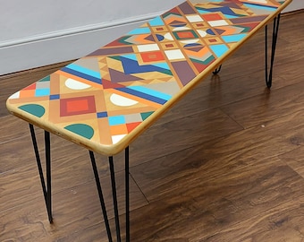 May include: A wooden bench with hairpin legs and a geometric pattern in shades of orange, blue, green, and brown.