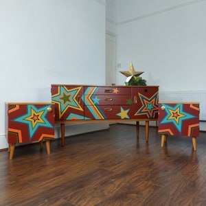 May include: A set of three mid-century modern side tables with a colourful geometric star pattern painted on the front. The tables are made of wood and have a brown stain. The largest table has three drawers and the two smaller tables have one drawer each.