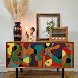 Puede incluir: Un aparador moderno de mediados de siglo con tres cajones y un diseño abstracto geométrico en tonos de verde, azul, amarillo, rojo y negro. El aparador tiene tiradores de metal negro y se asienta sobre cuatro patas negras. Una radio vintage y una lámpara con una figurita de cerámica están encima del aparador.
