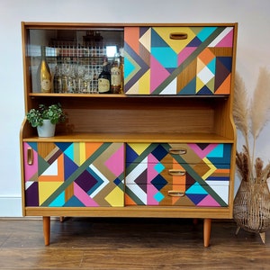 May include: A mid-century modern cabinet with a geometric pattern in shades of yellow, pink, blue, green, and brown. The cabinet has a glass top with a shelf and a lower section with two doors and drawers.