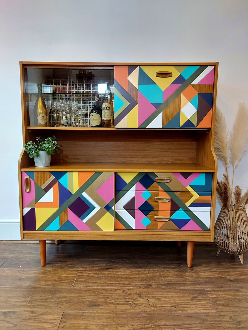 May include: A mid-century modern cabinet with a geometric pattern in shades of yellow, pink, blue, green, and brown. The cabinet has a glass top with a shelf and a lower section with two doors and drawers.
