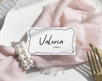 May include: A white place card with a black border and the name "Valeria" written in cursive. The place card is on a white plate with a pink napkin and pearl napkin ring. The table setting is complete with a gold fork and knife.