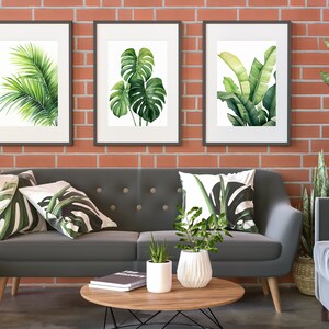 K&ouml;nnte beinhalten: Ein graues Sofa mit drei gr&uuml;nen Kissen mit Blattmotiven steht vor einer Backsteinwand mit drei gerahmten Drucken von tropischen Bl&auml;ttern. Eine Topfpflanze steht auf einem Couchtisch vor dem Sofa.