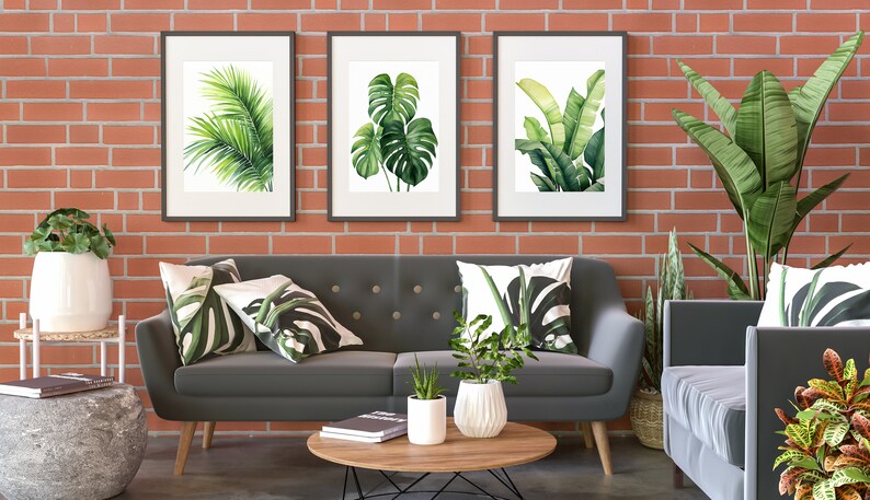 K&ouml;nnte beinhalten: Ein graues Sofa mit drei gr&uuml;nen Kissen mit Blattmotiven steht vor einer Backsteinwand mit drei gerahmten Drucken von tropischen Bl&auml;ttern. Eine Topfpflanze steht auf einem Couchtisch vor dem Sofa.