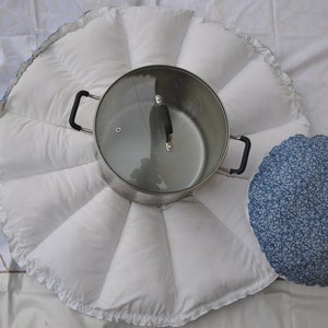 Peut inclure: Un coussin rond matelassé blanc avec un bord festonné. Le coussin a huit sections et une casserole en acier inoxydable avec un couvercle en verre au centre. Un coussin à motif floral bleu et blanc est à droite de la casserole.