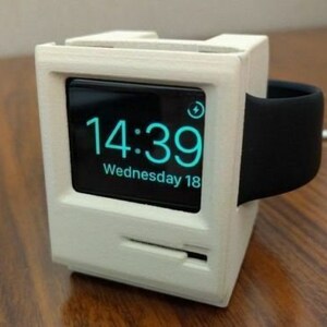 Peut inclure: Un support blanc imprimé en 3D en forme de vieux Macintosh avec une Apple Watch noire en charge à l'intérieur. Le cadran de la montre affiche l'heure 14:39 et la date mercredi 18.