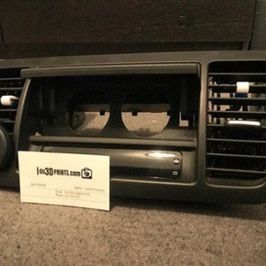 May include: A black plastic car dashboard trim piece with two round openings and a rectangular opening for a radio. The trim piece has two air vents on the sides. The trim piece is sitting on a brown carpet.