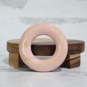 Op de afbeelding: Een roze keramische ring met een licht getextureerd oppervlak. De ring is dik en heeft een groot gat in het midden.