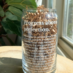 May include: A clear glass cup with a white inscription that reads "Progress above perfection. What can I be curious about today? A growth mindset is committed to learning rather than fearing the unknown. For you grow so much more by trying, whether you feel ready or not. Consistency in your actions, even if they are small, yields a far more beautiful journey. Keep going, my love. You are resilient. You are bright. You are abundant with divine purpose. There is no need to fear, dear one. You are exactly where you need to be. And you have everything you need for this journey."