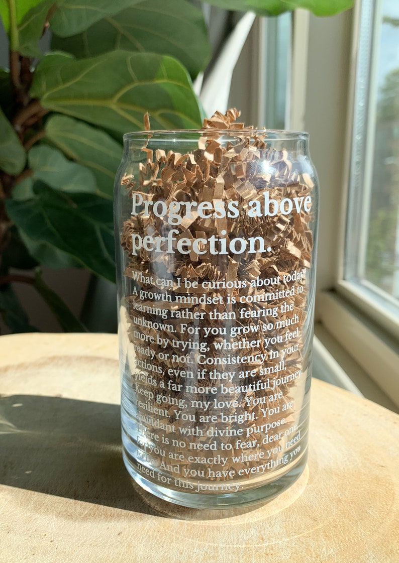 May include: A clear glass cup with a white inscription that reads "Progress above perfection. What can I be curious about today? A growth mindset is committed to learning rather than fearing the unknown. For you grow so much more by trying, whether you feel ready or not. Consistency in your actions, even if they are small, yields a far more beautiful journey. Keep going, my love. You are resilient. You are bright. You are abundant with divine purpose. There is no need to fear, dear one. You are exactly where you need to be. And you have everything you need for this journey."