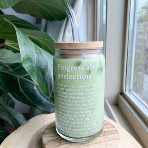 May include: A glass jar with a bamboo lid and a glass straw. The jar contains a green liquid and has a white label with the text "Progress above perfection." The text on the label reads: "What can I be curious about today! A growth mindset is committed to learning rather than fearing the unknown. For you grow so much more by trying, whether you feel ready or not. Consistency in your actions, even if they are small, yields a not. Consistency in your actions, even if they are small, yields a far more beautiful journey. Keep going, my love. You are resilient. You are bright. You are abundant with divine purpose. There is no need to fear, dear one. For you are exactly where you need to be. And you have everything you need for this journey."