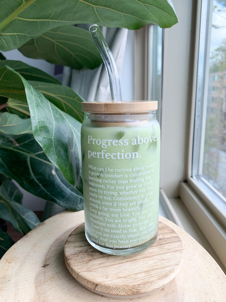 May include: A glass jar with a bamboo lid and a glass straw. The jar contains a green liquid and has a white label with the text "Progress above perfection." The text on the label reads: "What can I be curious about today! A growth mindset is committed to learning rather than fearing the unknown. For you grow so much more by trying, whether you feel ready or not. Consistency in your actions, even if they are small, yields a not. Consistency in your actions, even if they are small, yields a far more beautiful journey. Keep going, my love. You are resilient. You are bright. You are abundant with divine purpose. There is no need to fear, dear one. For you are exactly where you need to be. And you have everything you need for this journey."