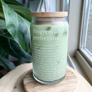 May include: A glass jar with a bamboo lid filled with a green liquid. The jar has a white label with the text "Progress above perfection." and a quote about growth mindset.