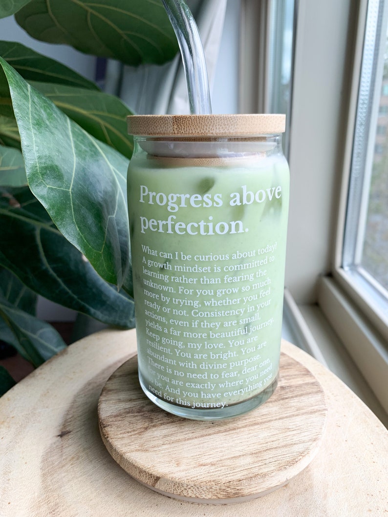 May include: A glass jar with a bamboo lid filled with a green liquid. The jar has a white label with the text "Progress above perfection." and a quote about growth mindset.