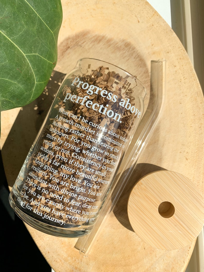 May include: A clear glass bottle with a white inscription that reads "Progress above perfection. What can I be curious about today? A growth mindset is committed to learning rather than fearing the unknown. For you grow so much more by trying, whether you feel ready or not. Consistency in your actions, even if they are small, yields a far more beautiful journey. Keep going, my love. You are resilient. You are bright. You are abundant with divine purpose. There is no need to fear, dear one. You are exactly where you need to be. And you have everything you need for this journey."