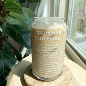 May include: A glass can with a brown beverage inside. The can has a white label with the text "Progress above perfection." and a quote about growth mindset and curiosity.