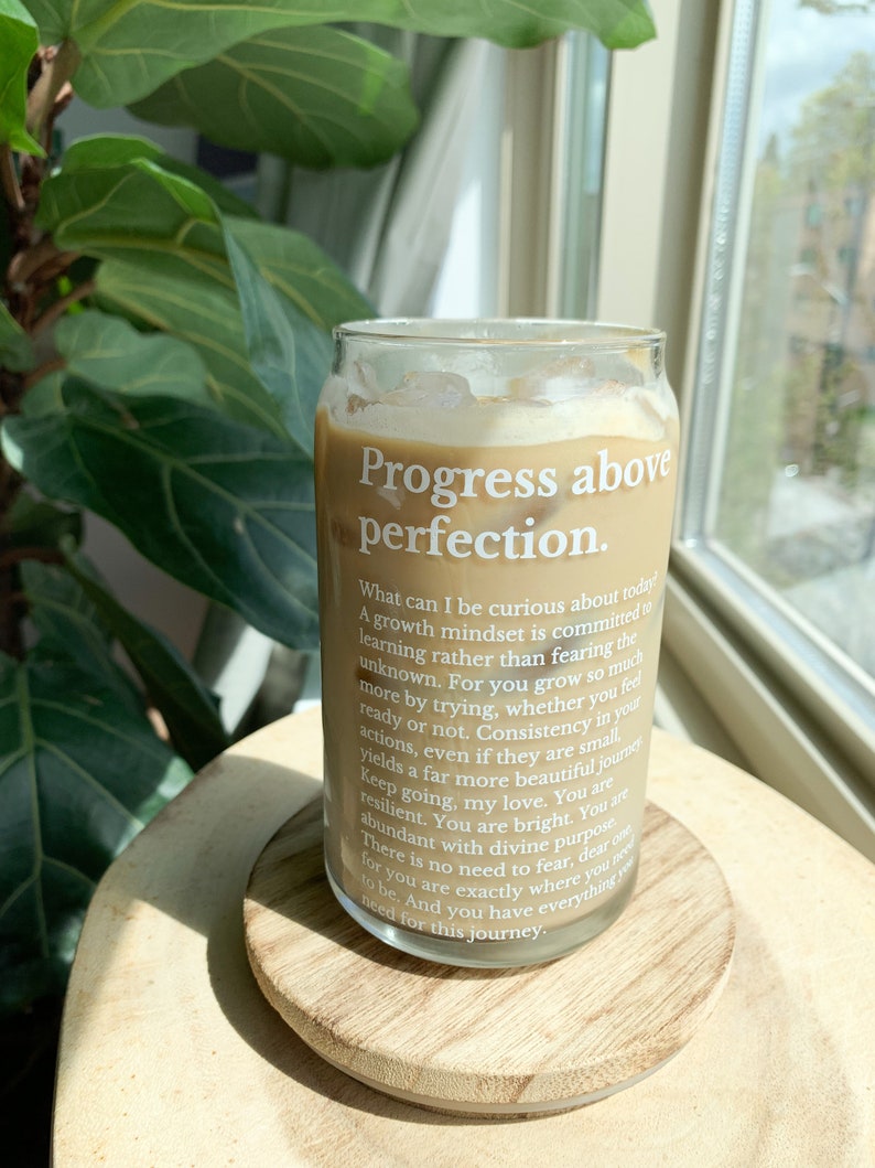 May include: A glass can with a brown beverage inside. The can has a white label with the text "Progress above perfection." and a quote about growth mindset and curiosity.