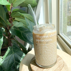 May include: A glass of iced coffee with a glass straw in a clear glass with the text "Progress above perfection."  The text continues with a motivational quote about growth mindset and consistency. The glass is sitting on a wooden coaster.