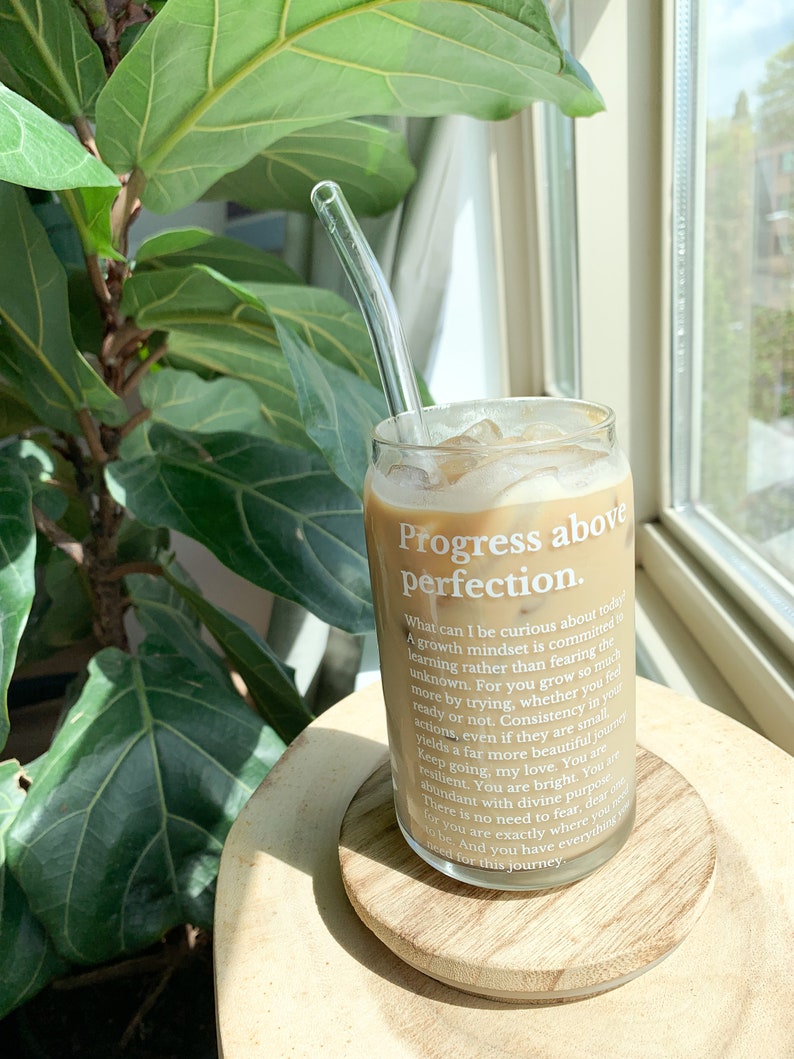 May include: A glass of iced coffee with a glass straw in a clear glass with the text "Progress above perfection."  The text continues with a motivational quote about growth mindset and consistency. The glass is sitting on a wooden coaster.