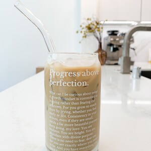 May include: A glass of iced coffee with a glass straw in a clear glass with the text "Progress above perfection." and a motivational quote about growth mindset.