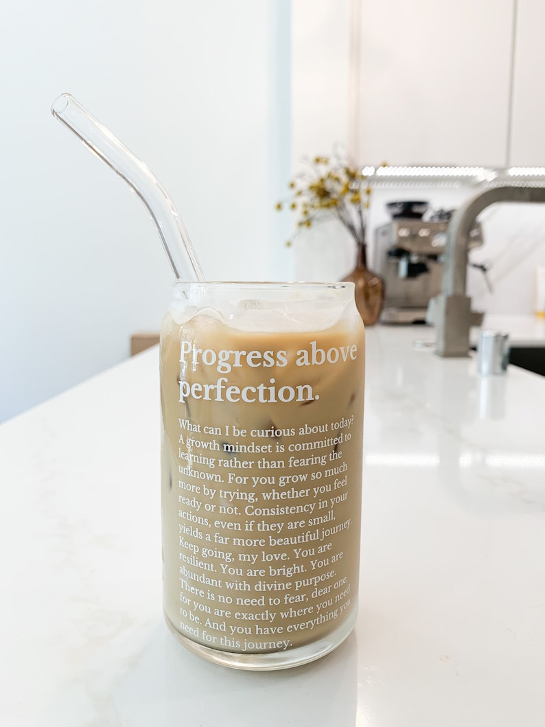 May include: A glass of iced coffee with a glass straw in a clear glass with the text "Progress above perfection." and a motivational quote about growth mindset.