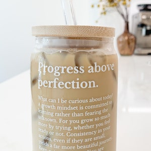 May include: A glass jar with a bamboo lid filled with iced coffee and a glass straw. The jar has a white label with the text "Progress above perfection." and a quote about growth mindset.