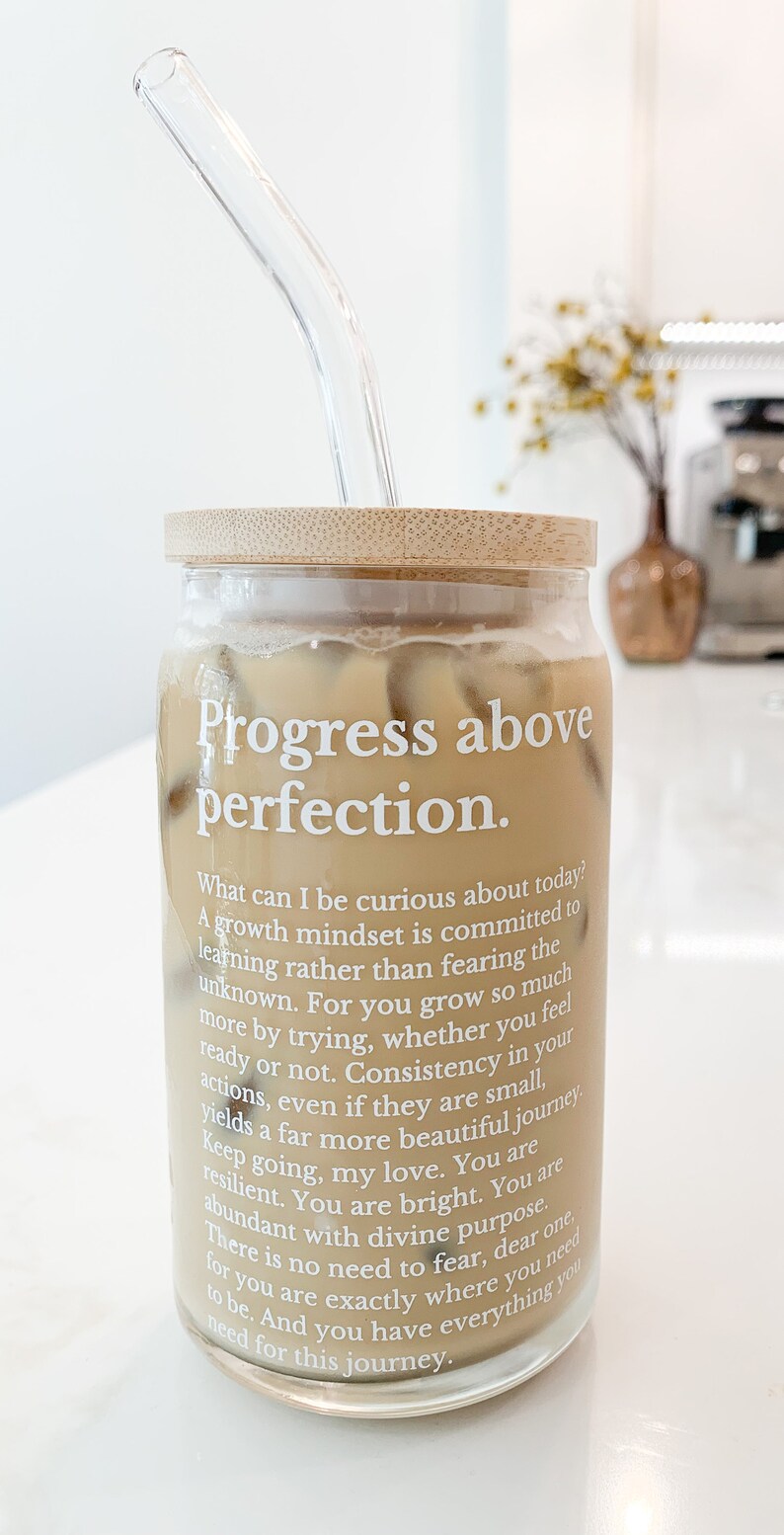 May include: A glass jar with a bamboo lid filled with iced coffee and a glass straw. The jar has a white label with the text "Progress above perfection." and a quote about growth mindset.