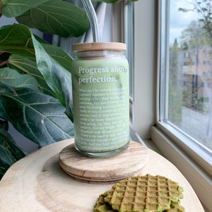 May include: A glass jar with a bamboo lid and a glass straw. The jar is filled with a green liquid and has the text "Progress above perfection." printed on it. The jar is sitting on a wooden surface with two green waffles.