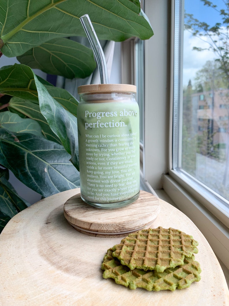May include: A glass jar with a bamboo lid and a glass straw. The jar is filled with a green liquid and has the text "Progress above perfection." printed on it. The jar is sitting on a wooden surface with two green waffles.