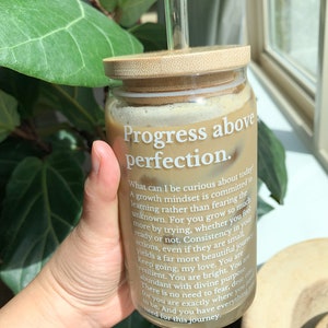 May include: A glass jar with a bamboo lid and a glass straw filled with a brown liquid. The jar has a white label with the text "Progress above perfection." and a motivational quote about growth mindset.