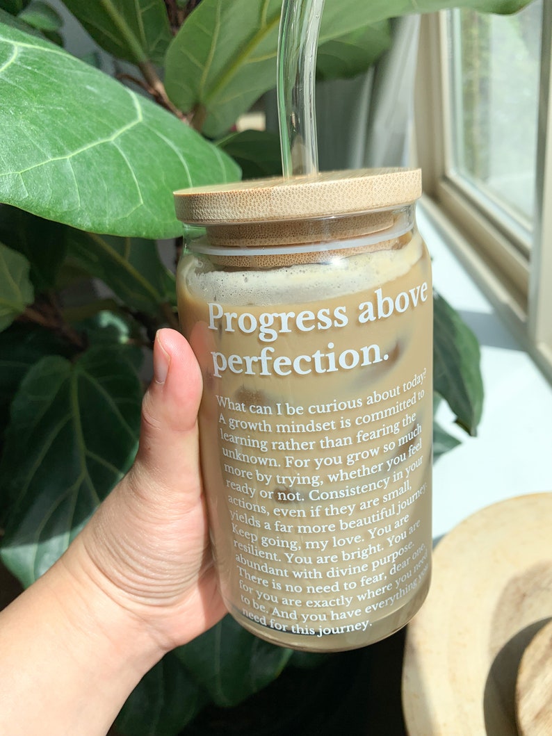 May include: A glass jar with a bamboo lid and a glass straw filled with a brown liquid. The jar has a white label with the text "Progress above perfection." and a motivational quote about growth mindset.
