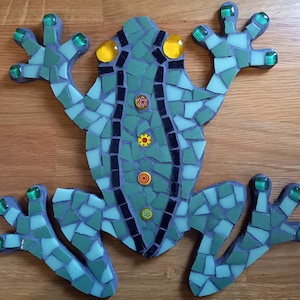 Op de afbeelding: Een mozaïek kikker gemaakt van groene, blauwe, zwarte en gele tegels. De kikker heeft een zwarte streep op zijn rug en vier gele bloemen op zijn lichaam.