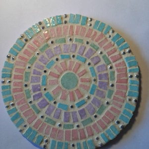 Peut inclure: Un dessous de verre rond en mosaïque avec un motif circulaire dans des tons de rose, bleu, vert et violet. Le centre du dessous de verre présente un cercle plus petit de carreaux vert clair.