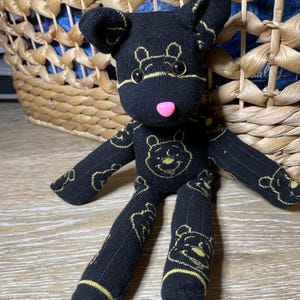 Puede incluir: Un oso de calcetín negro hecho a mano con gráficos amarillos de Winnie the Pooh. El oso tiene una nariz rosa y ojos negros de botón. El oso está frente a una cesta de mimbre.