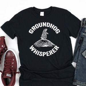 May include: A black t-shirt with a white graphic of a groundhog standing on a mound of dirt. The graphic is surrounded by a circle with the text "Groundhog Whisperer". The shirt is paired with dark blue jeans, a silver watch, and maroon high-top sneakers.