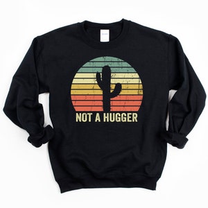 Puede incluir: Sudadera negra con un gr&aacute;fico vintage de un cactus en una puesta de sol. El texto "NOT A HUGGER" est&aacute; impreso debajo del cactus.