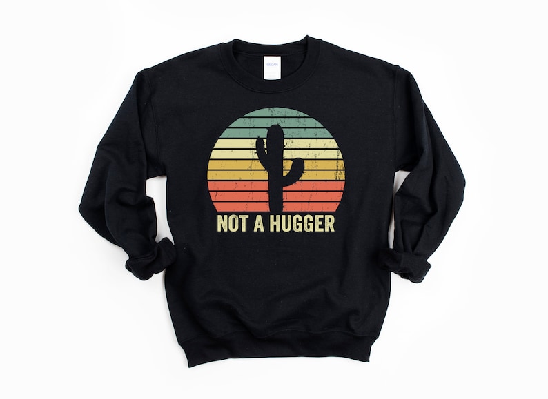 Puede incluir: Sudadera negra con un gr&aacute;fico vintage de un cactus en una puesta de sol. El texto "NOT A HUGGER" est&aacute; impreso debajo del cactus.