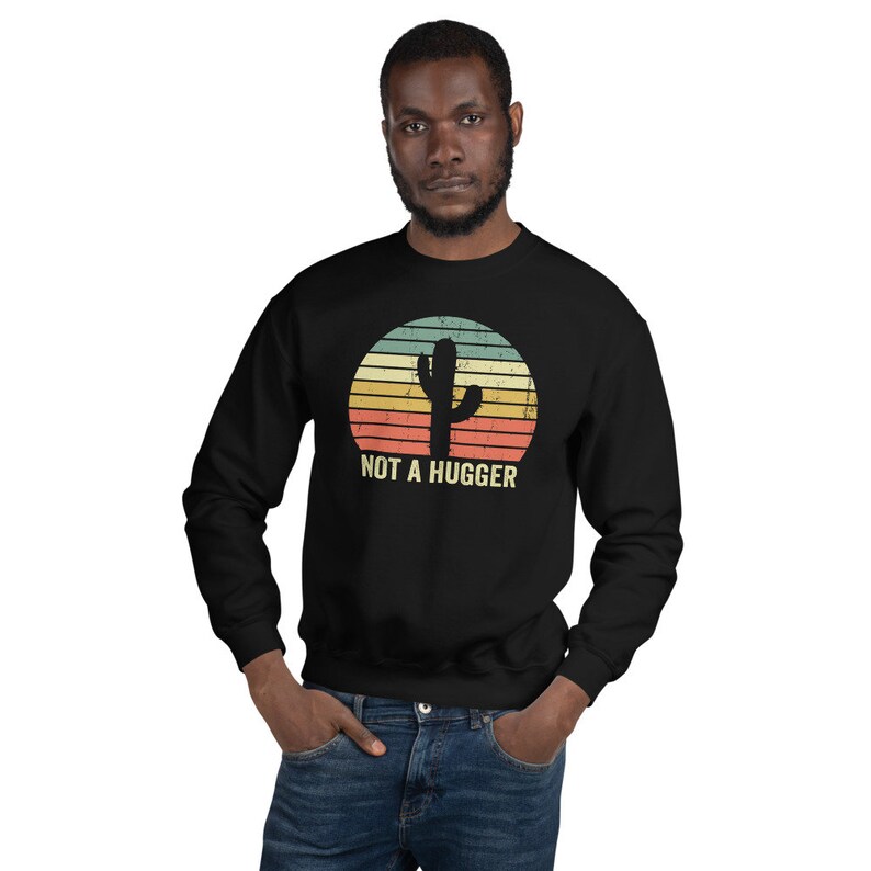 Puede incluir: Sudadera negra con un gr&aacute;fico vintage de un cactus en una puesta de sol. El texto "NOT A HUGGER" est&aacute; impreso debajo del cactus.