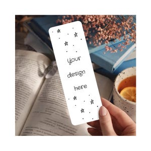 May include: A white bookmark with black star and dot designs, and the text "your design here." The bookmark is held in a hand, with an open book and a cup of tea in the background.