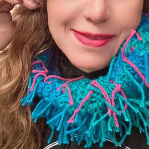 May include: A close-up of a blue and pink crocheted neck warmer with long fringe. The neck warmer is made with a soft, textured yarn.