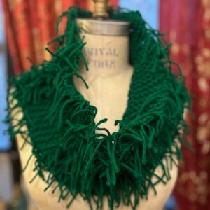 May include: A green knitted cowl with long fringe. The cowl is displayed on a mannequin.