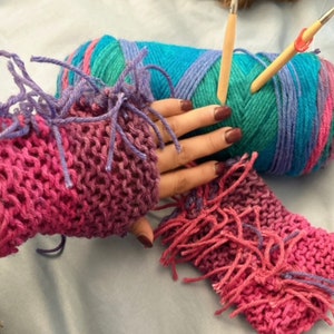 May include: A pair of pink and purple knitted wrist warmers with fringe. The wrist warmers are made with a chunky yarn and have a gradient color pattern. The wrist warmers are lying on a blue surface next to a ball of yarn and knitting needles.