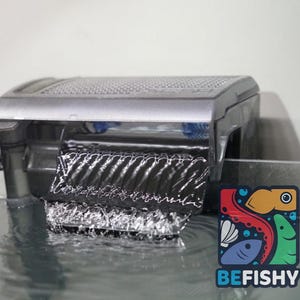 May include: A silver and black aquarium hang-on-back power filter with a clear plastic water flow tray, featuring a textured top lid and a visible water output stream cascading into the tank, with a BEFISHY logo in the corner.