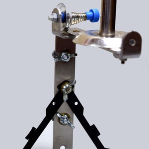 Puede incluir: Una herramienta de metal plateado con un resorte y una perilla azul. La herramienta tiene tres patas negras que se extienden desde la base. La herramienta está diseñada para usarse con una máquina de coser.