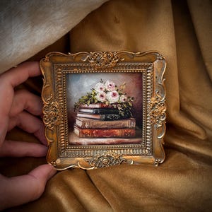 May include: A small, framed painting featuring a stack of books topped with a bouquet of pink and white roses. The frame is ornate, gold-colored, and has a detailed floral design. The painting has a vintage aesthetic.