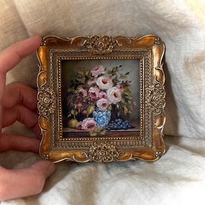 May include: A framed oil painting of a still life featuring a bouquet of pink roses in a blue and white porcelain vase. The vase is surrounded by grapes, a pear, and green leaves. The painting is set in a gold ornate frame.