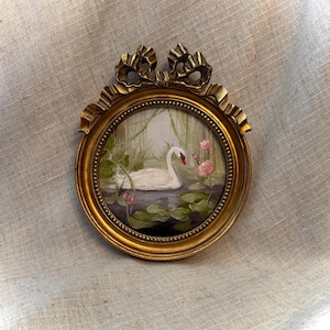 May include: A circular painting of a white swan swimming in a pond with green lily pads and pink flowers, encased in a gold-toned frame featuring a decorative bow at the top. The artwork depicts a serene nature scene with soft brushstrokes.