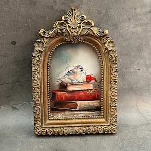 Mini lámina artística enmarcada con la imagen de un pájaro sobre una pila de libros.